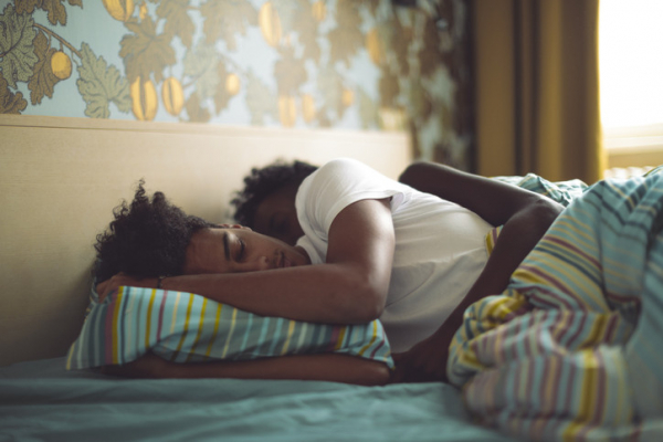Couple asleep in bed with multicolored striped pillow case and quilt, morning light coming through window; one has arm over the other